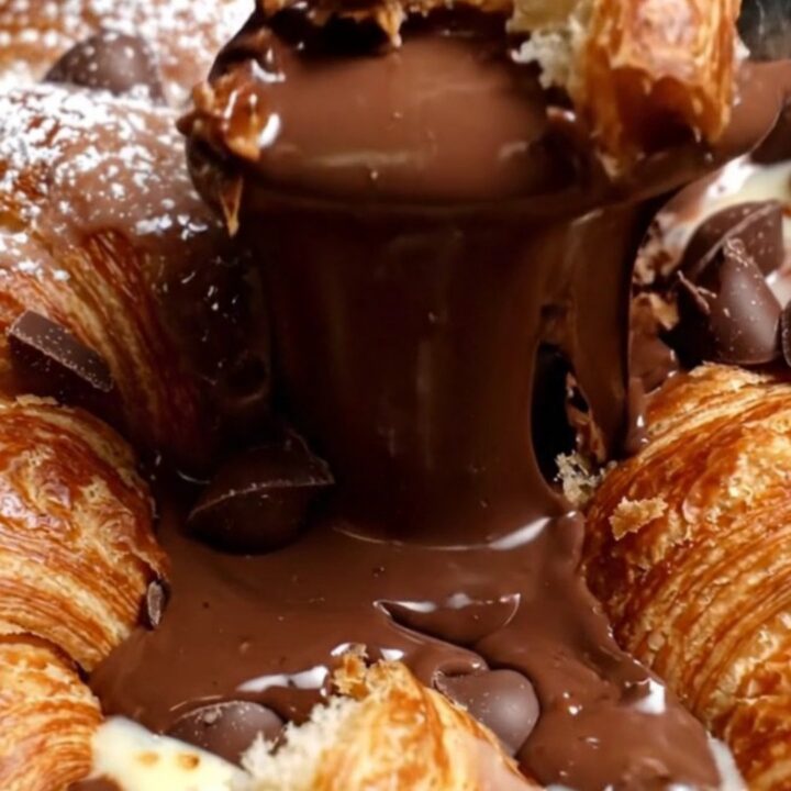 A close-up of a spoon breaking the crispy, golden-brown crust of a croissant chocolate pudding skillet.