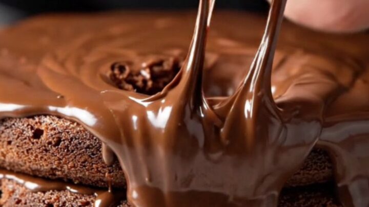 A towering three-layer chocolate cake with glossy fudge frosting and a fork sinking into it.