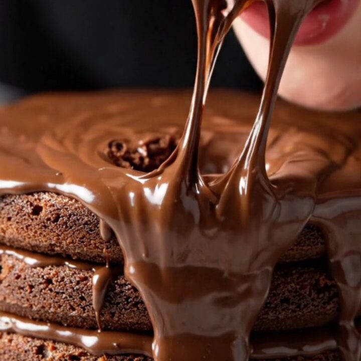 A towering three-layer chocolate cake with glossy fudge frosting and a fork sinking into it.