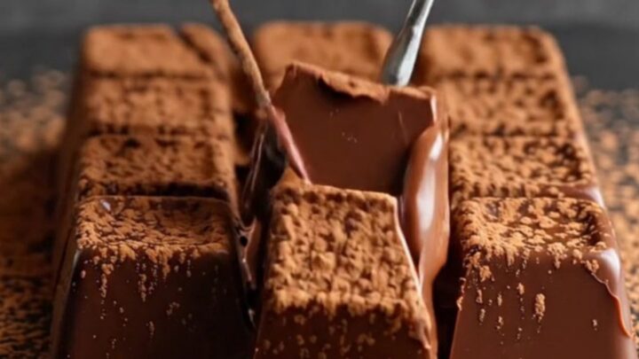 A close-up of a warm knife slicing through a velvety block of dark chocolate dusted with cocoa powder.