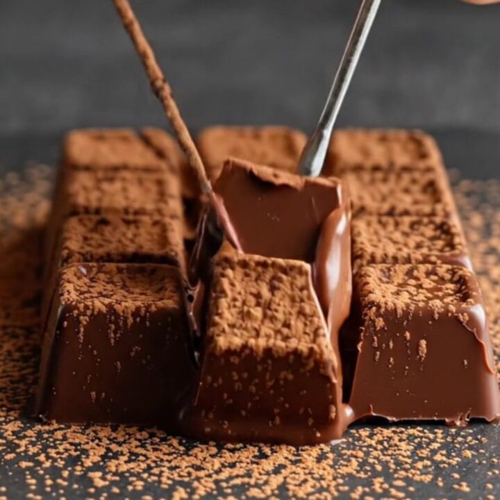 A close-up of a warm knife slicing through a velvety block of dark chocolate dusted with cocoa powder.