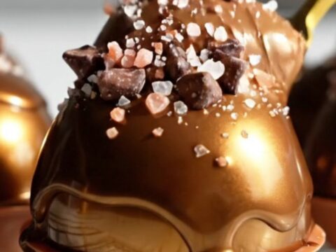 A close-up shot of a gold spoon scooping into a glossy, dark chocolate mousse dome.