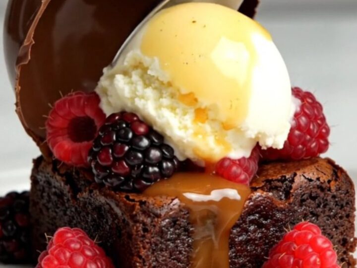 A glossy chocolate dome sitting on a white plate with a brownie, gelato, and berries.
