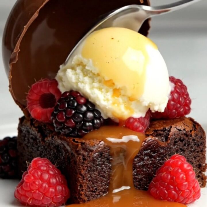 A glossy chocolate dome sitting on a white plate with a brownie, gelato, and berries.