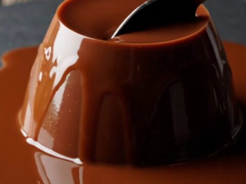 A close-up of a glossy, dark chocolate espresso panna cotta dome on a dark slate plate with a spoon resting nearby.