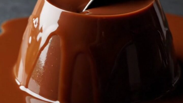 A close-up of a glossy, dark chocolate espresso panna cotta dome on a dark slate plate with a spoon resting nearby.