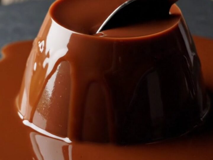 A close-up of a glossy, dark chocolate espresso panna cotta dome on a dark slate plate with a spoon resting nearby.
