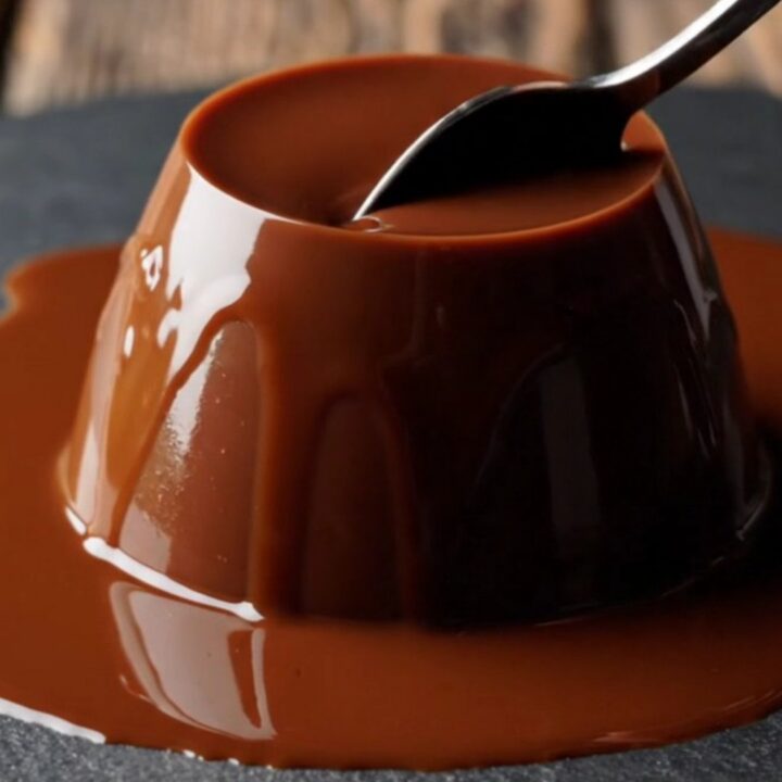 A close-up of a glossy, dark chocolate espresso panna cotta dome on a dark slate plate with a spoon resting nearby.