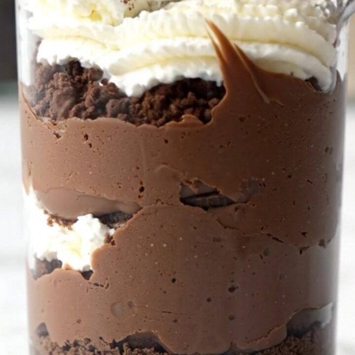 A close-up shot of a glass cup filled with layers of crushed Oreos, dark chocolate pudding, and glossy ganache.