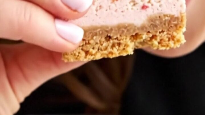 A close-up of a strawberry crunch cheesecake bar showing layers of golden crust, creamy vanilla filling