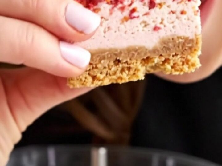 A close-up of a strawberry crunch cheesecake bar showing layers of golden crust, creamy vanilla filling