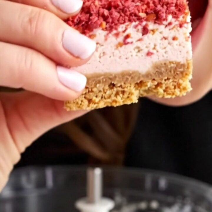 A close-up of a strawberry crunch cheesecake bar showing layers of golden crust, creamy vanilla filling
