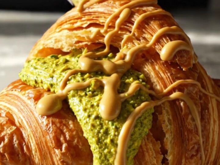 A close-up of a golden, circular Suprême croissant with vibrant green pistachio cream bursting from the center.
