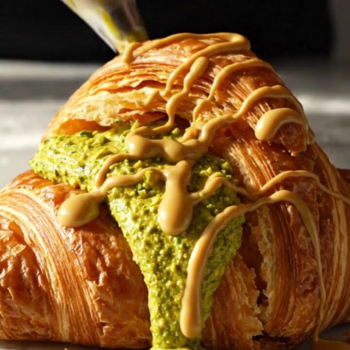 A close-up of a golden, circular Suprême croissant with vibrant green pistachio cream bursting from the center.
