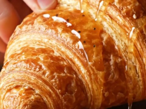 A golden, flaky everything bagel croissant drizzled with hot honey, showing a glimpse of the creamy filling inside.