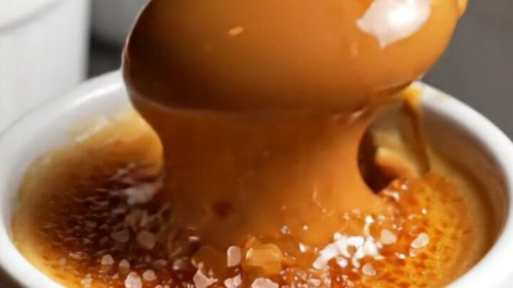 A close-up of a spoon cracking the golden-brown caramelized sugar shell of a dulce de leche crème brûlée.