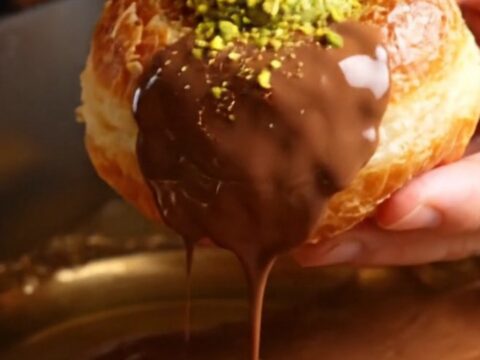 A close-up of a golden fried brioche doughnut split open to reveal a vibrant green pistachio and kataifi filling.