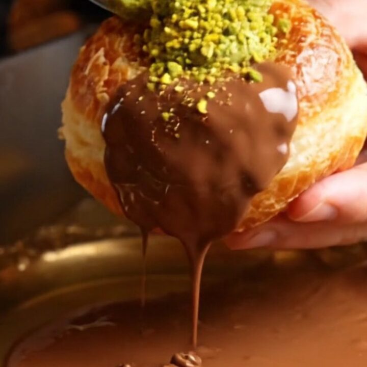 A close-up of a golden fried brioche doughnut split open to reveal a vibrant green pistachio and kataifi filling.