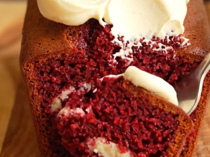 A moist red velvet loaf cake with a cream cheese swirl, topped with thick vanilla frosting and cake crumbs.