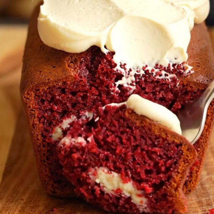 A moist red velvet loaf cake with a cream cheese swirl, topped with thick vanilla frosting and cake crumbs.