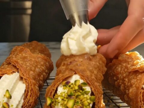A close-up of a freshly filled Sicilian cannoli with a golden, blistered shell and crushed pistachios on the ends.