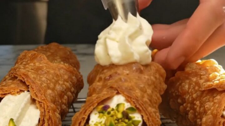 A close-up of a freshly filled Sicilian cannoli with a golden, blistered shell and crushed pistachios on the ends.