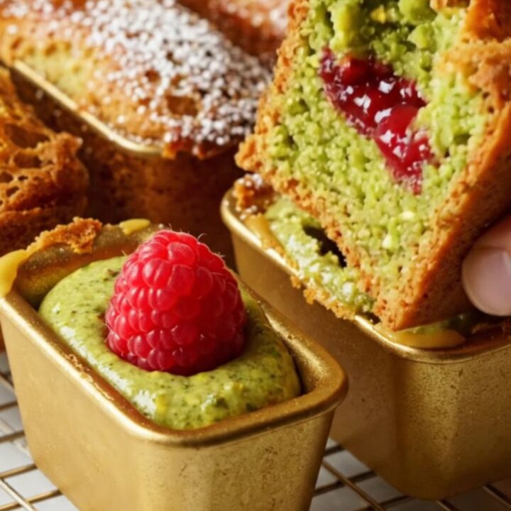 A golden brown pistachio financier broken in half to reveal a vibrant green crumb and a jammy raspberry center.