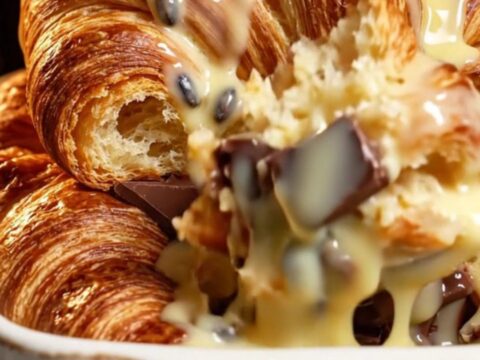 A close-up of a golden brown croissant bread pudding with melted dark chocolate and vanilla sauce.
