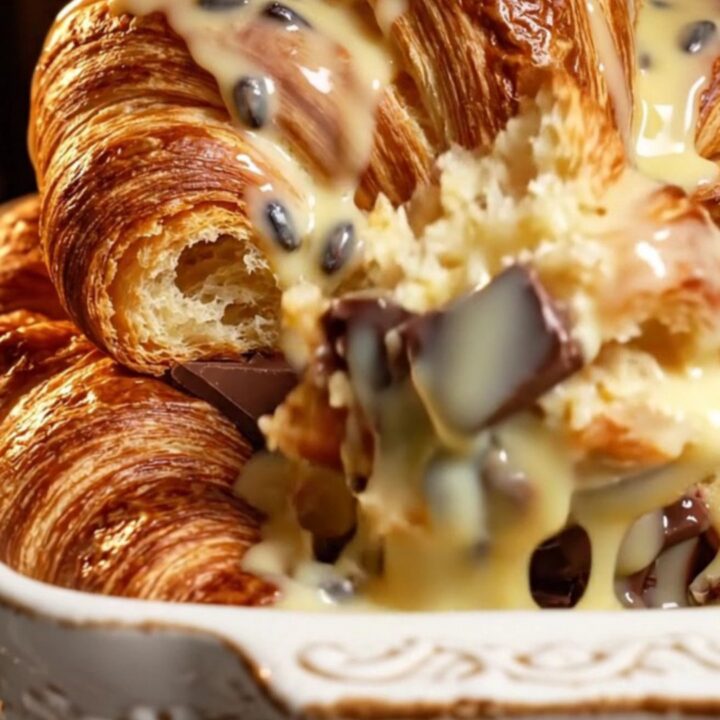 A close-up of a golden brown croissant bread pudding with melted dark chocolate and vanilla sauce.