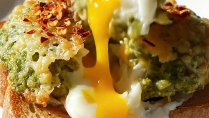 A close-up of two sunny-side-up eggs with a crispy Parmesan lace and green pesto, resting on a slice of toasted brioche.