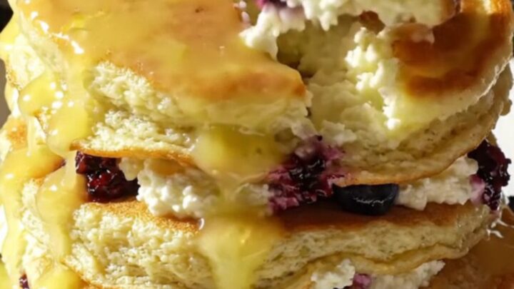 A tall stack of fluffy lemon ricotta pancakes topped with glossy yellow lemon curd and fresh blueberries.