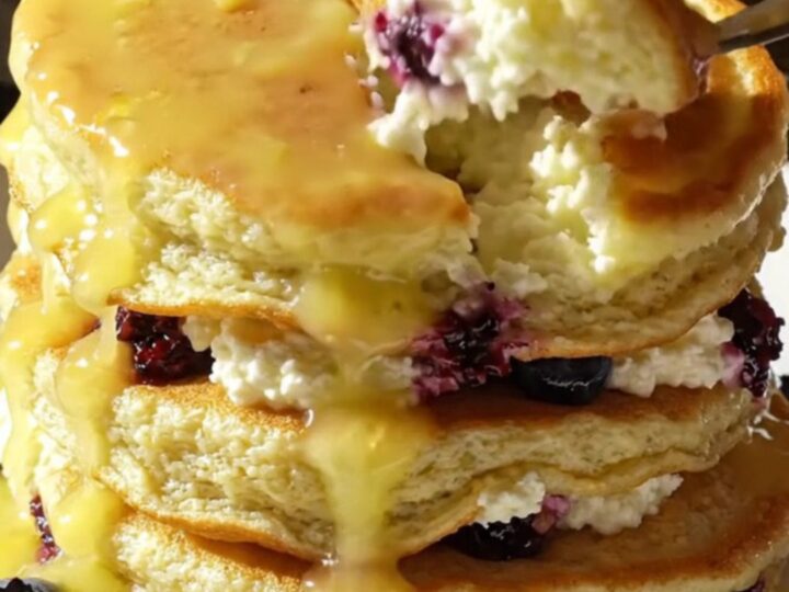 A tall stack of fluffy lemon ricotta pancakes topped with glossy yellow lemon curd and fresh blueberries.