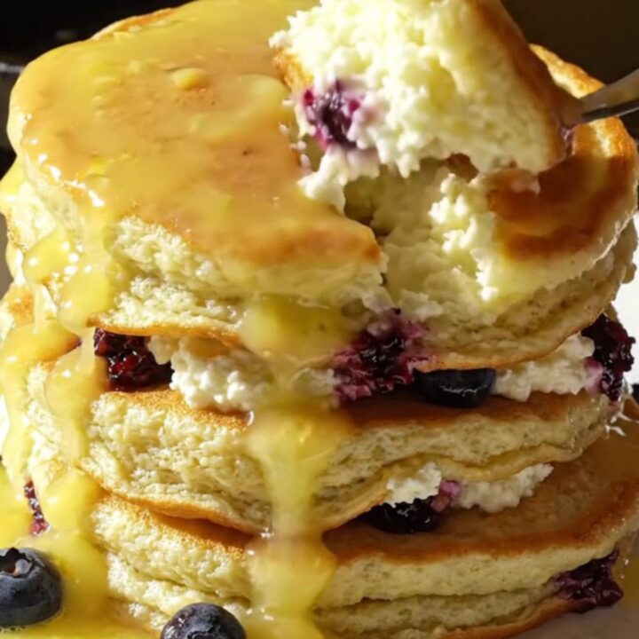 A tall stack of fluffy lemon ricotta pancakes topped with glossy yellow lemon curd and fresh blueberries.