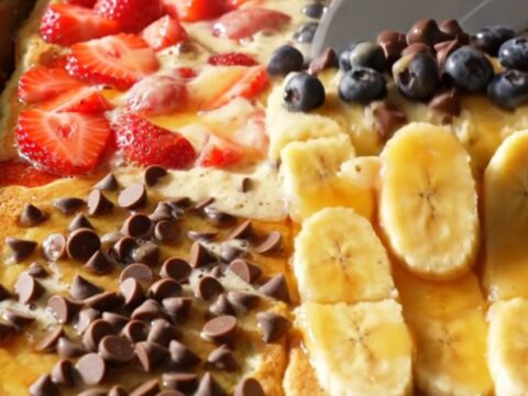 A golden, fluffy sheet pan pancake divided into four distinct quadrants featuring strawberries, blueberries, chocolate