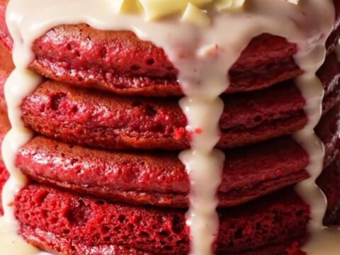 A stack of three vibrant crimson red velvet pancakes topped with a cascading ivory cream cheese glaze.