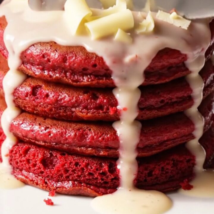 A stack of three vibrant crimson red velvet pancakes topped with a cascading ivory cream cheese glaze.