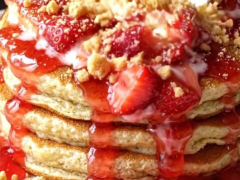 A stack of golden brown pancakes topped with fresh macerated strawberries and crushed graham crackers.