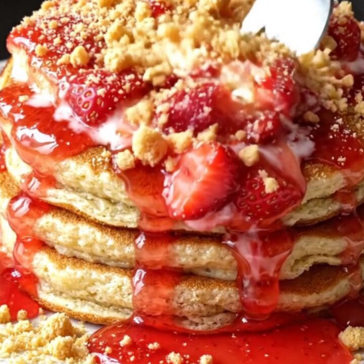A stack of golden brown pancakes topped with fresh macerated strawberries and crushed graham crackers.