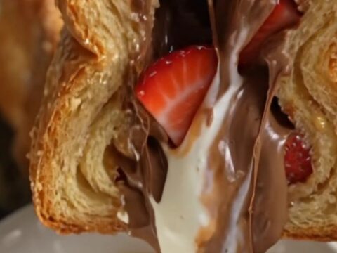 A close-up of a stuffed brioche French toast slice being pulled apart, revealing a molten Nutella and strawberry filling.