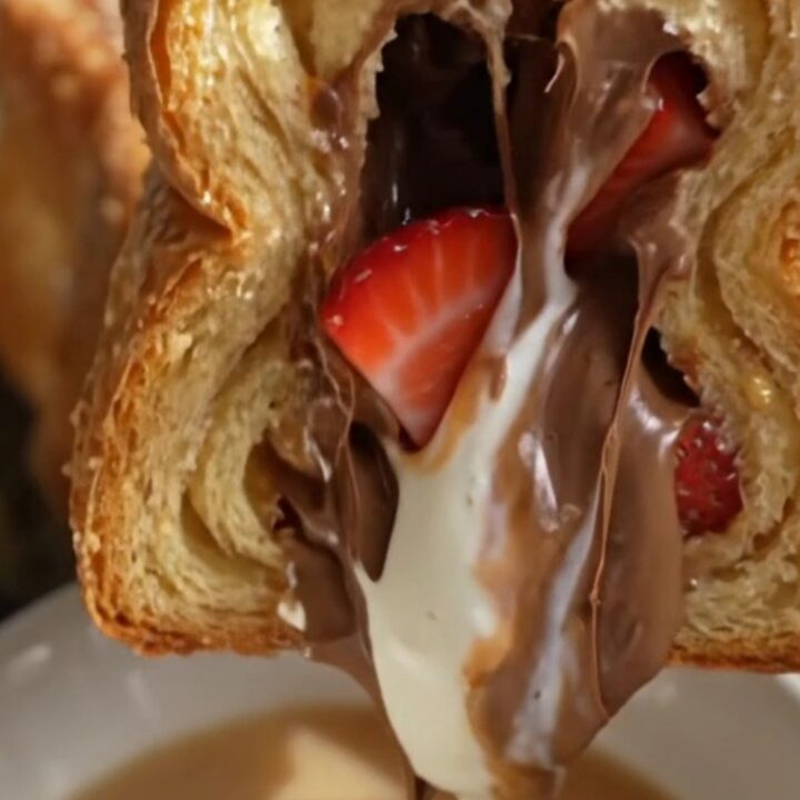 A close-up of a stuffed brioche French toast slice being pulled apart, revealing a molten Nutella and strawberry filling.