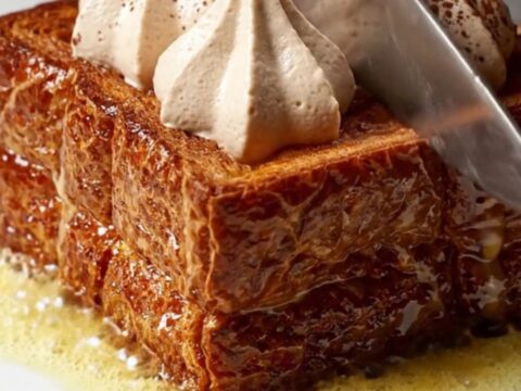 A close-up of thick, caramelized brioche French toast topped with piped swirls of mascarpone cream and dusted with cocoa
