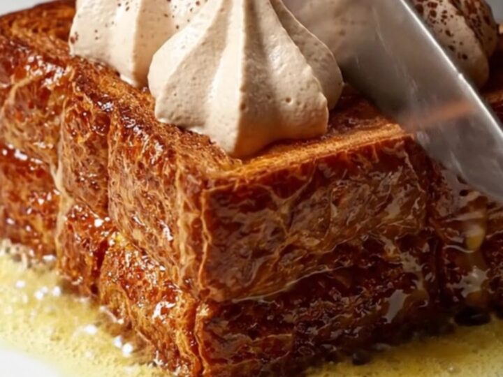 A close-up of thick, caramelized brioche French toast topped with piped swirls of mascarpone cream and dusted with cocoa