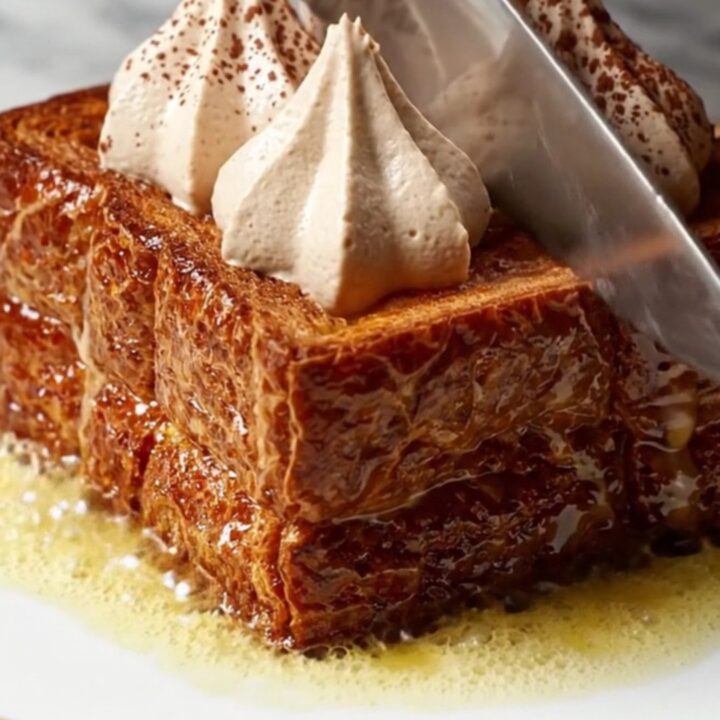 A close-up of thick, caramelized brioche French toast topped with piped swirls of mascarpone cream and dusted with cocoa