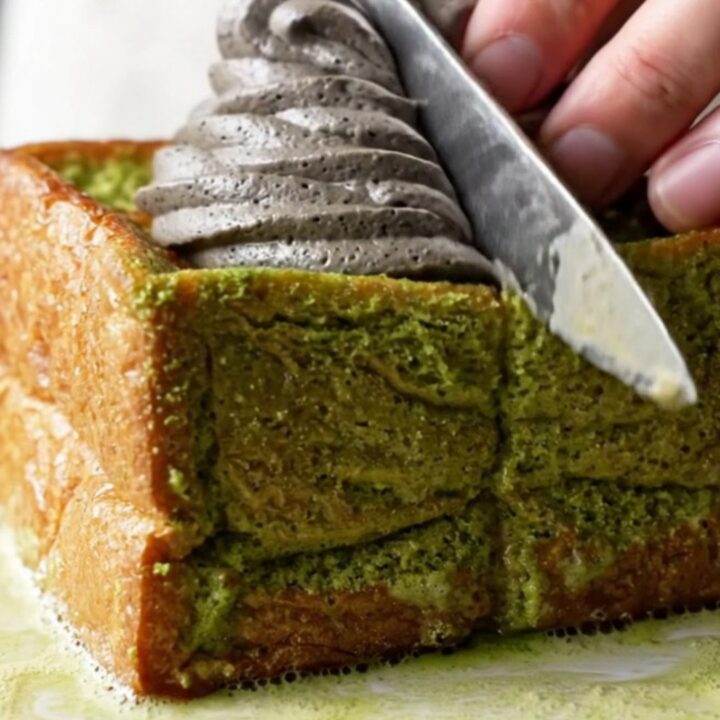 A thick square of matcha French toast with golden edges, topped with a dramatic swirl of black sesame whipped cream.