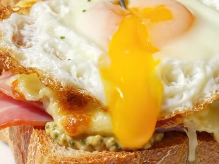 A golden-broiled Croque Madame topped with a sunny-side up egg on a marble bistro table.