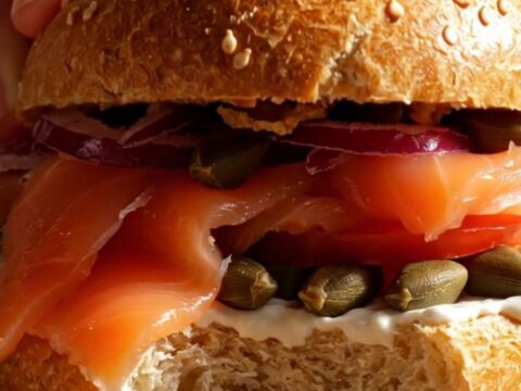 A close-up of a toasted everything bagel topped with creamy scallion-dill schmear, silky Nova lox, and fresh tomato slices.