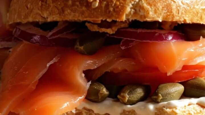 A close-up of a toasted everything bagel topped with creamy scallion-dill schmear, silky Nova lox, and fresh tomato slices.