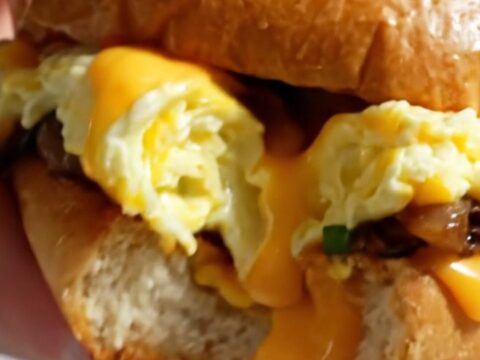 A close-up of a soft-scrambled egg sandwich on a toasted brioche bun with chives and visible melted cheddar.