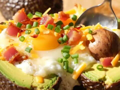 A close-up of a baked avocado half topped with a runny egg, melted cheddar, and crispy pancetta on a wooden board.