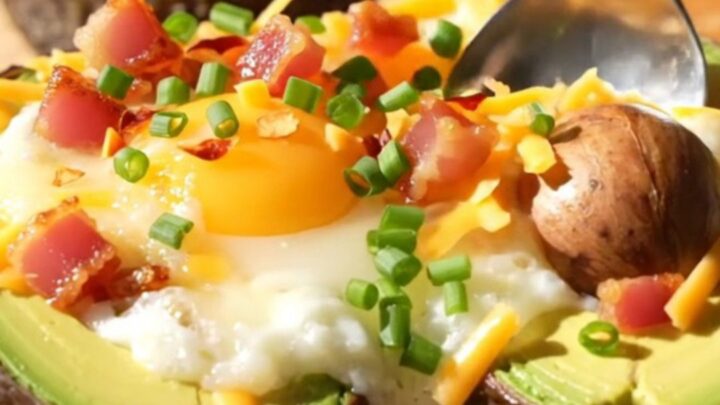 A close-up of a baked avocado half topped with a runny egg, melted cheddar, and crispy pancetta on a wooden board.